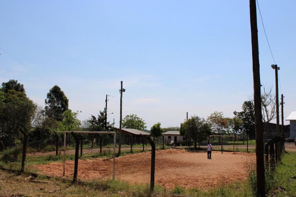 Foto colorida horizontal. À direita, na parte de baixo da foto, uma garota sozinha no meio do campo. Ela olha para o chão de areia. Ao redor, o campo está cercado por estacas de madeira, postes e árvores e grama.
