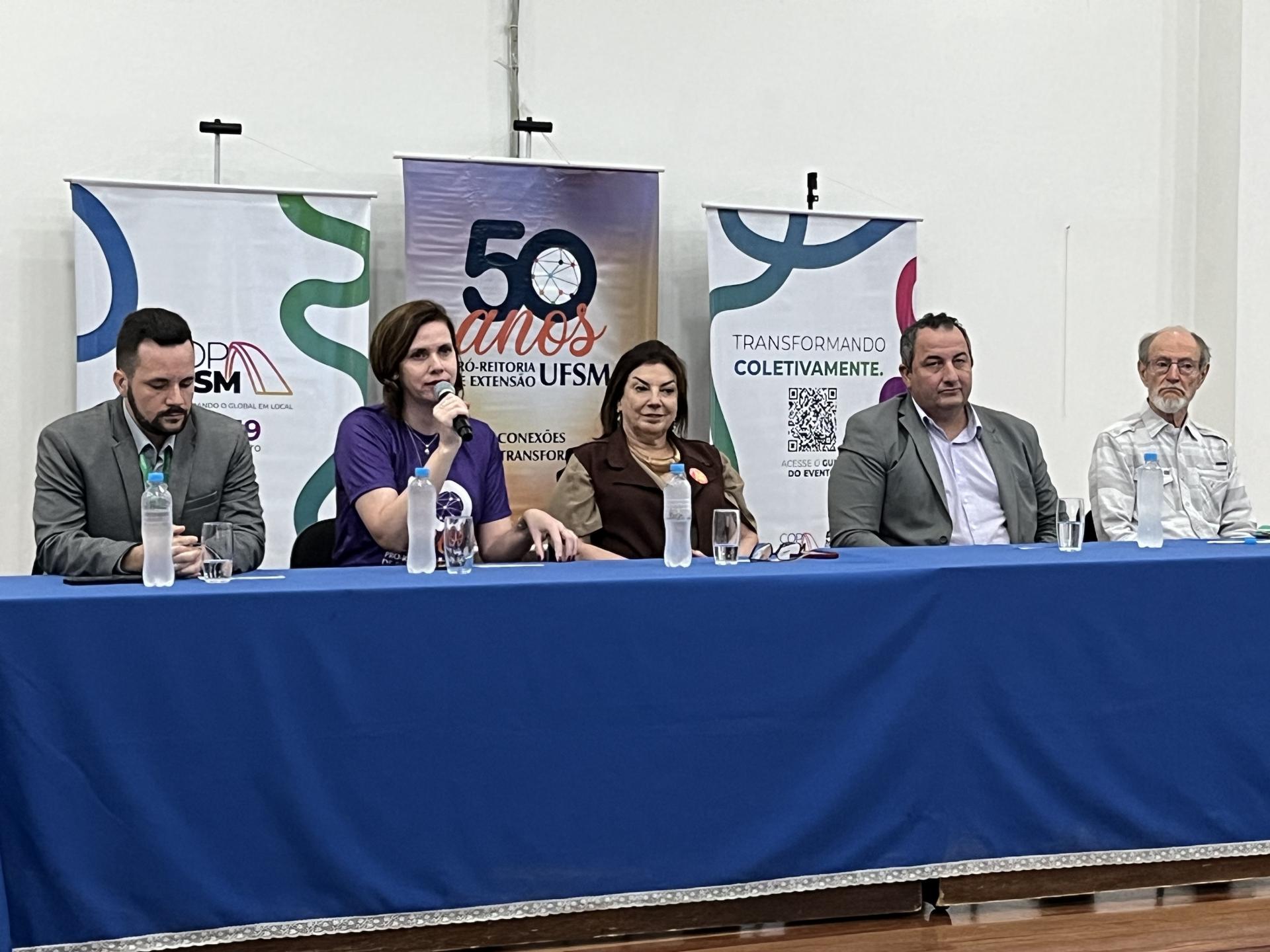 Cinco pessoas estão sentadas em uma mesa coberta por um pano azul; são três homens e duas mulheres. Na mesa, há garrafas de água transparentes e copos de vidro. Atrás, há uma parede branca, e três banners coloridos sobre a COP UFSM e os 50 anos da Pró-Reitoria de Extensão.