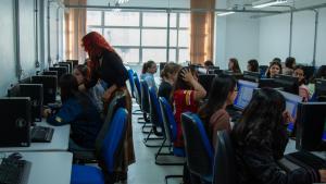 Um grupo de alunas está em uma sala. Ao fundo, há uma janela com cortinas. A sala tem várias mesas com computadores, e várias alunas sentadas. Uma mulher está de pé, ajudando uma das alunas.