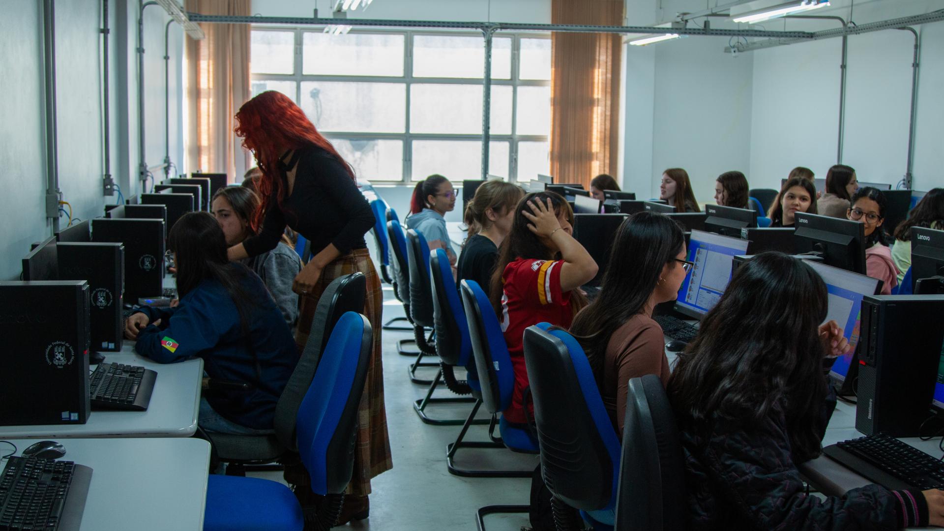 Um grupo de alunas está em uma sala. Ao fundo, há uma janela com cortinas. A sala tem várias mesas com computadores, e várias alunas sentadas. Uma mulher está de pé, ajudando uma das alunas.