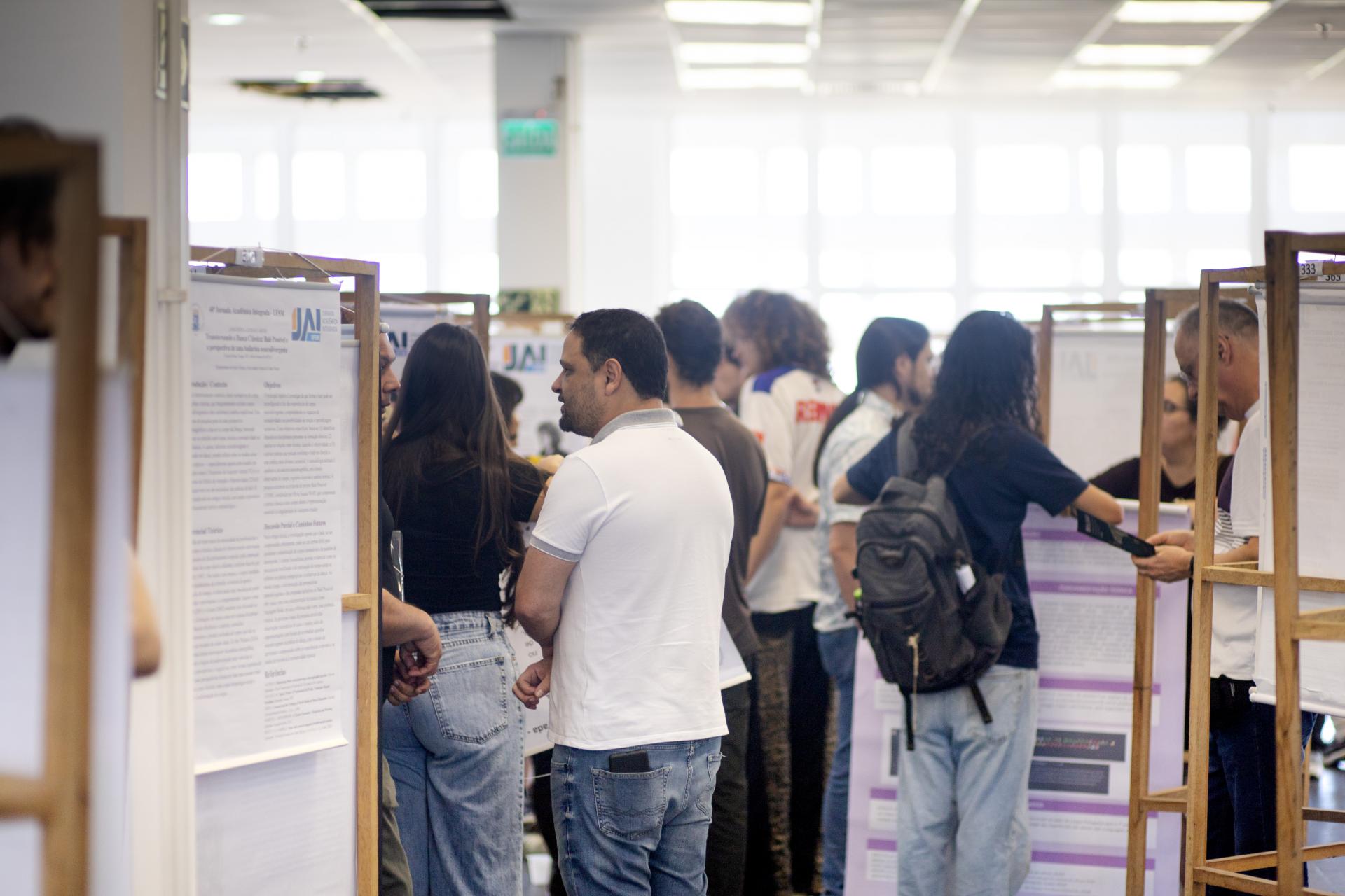 Foto horizontal colorida. Mostra o ambiente interno do Centro de Convenções movimentado, com pessoas apresentando pôsteres acadêmicos. Em primeiro plano, pessoas conversam e observam os pôsteres dispostos em suportes de madeira; a maioria veste roupas casuais, e algumas seguram pranchetas ou pastas. Ao fundo, o espaço amplo e bem iluminado indica um ambiente universitário, com janelas grandes e luz natural. À esquerda, vê-se parte de um pôster e o ombro de uma pessoa; ao centro, duas pessoas conversam em frente aos painéis; à direita, vários grupos observam os trabalhos, com cartazes da Jornada Acadêmica Integrada (JAI) visíveis.