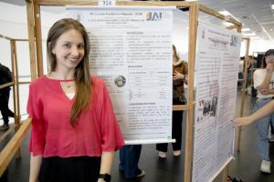 Foto horizontal colorida. Mostra a estudante Gabriele apresentando seu pôster durante a Jornada Acadêmica Integrada da UFSM. Em primeiro plano, a jovem sorri diante de seu pôster, que trata de um estudo sobre o “Jogo de imagens sobre o réu no Tribunal do Júri do Caso Kiss”. Atrás dela há outros pôsteres e pessoas circulando. O ambiente é bem iluminado, com estrutura de madeira sustentando os trabalhos expostos. À esquerda, a apresentadora e seu pôster; ao centro, os detalhes do texto e imagens do painel; à direita, outra pessoa observa um pôster vizinho.