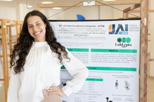 Foto horizontal colorida. Mostra o ambiente interno do Centro de Convenções movimentado, com pessoas apresentando pôsteres acadêmicos. Em primeiro plano, pessoas conversam e observam os pôsteres dispostos em suportes de madeira; a maioria veste roupas casuais, e algumas seguram pranchetas ou pastas. Ao fundo, o espaço amplo e bem iluminado indica um ambiente universitário, com janelas grandes e luz natural. À esquerda, vê-se parte de um pôster e o ombro de uma pessoa; ao centro, duas pessoas conversam em frente aos painéis; à direita, vários grupos observam os trabalhos, com cartazes da Jornada Acadêmica Integrada (JAI) visíveis.