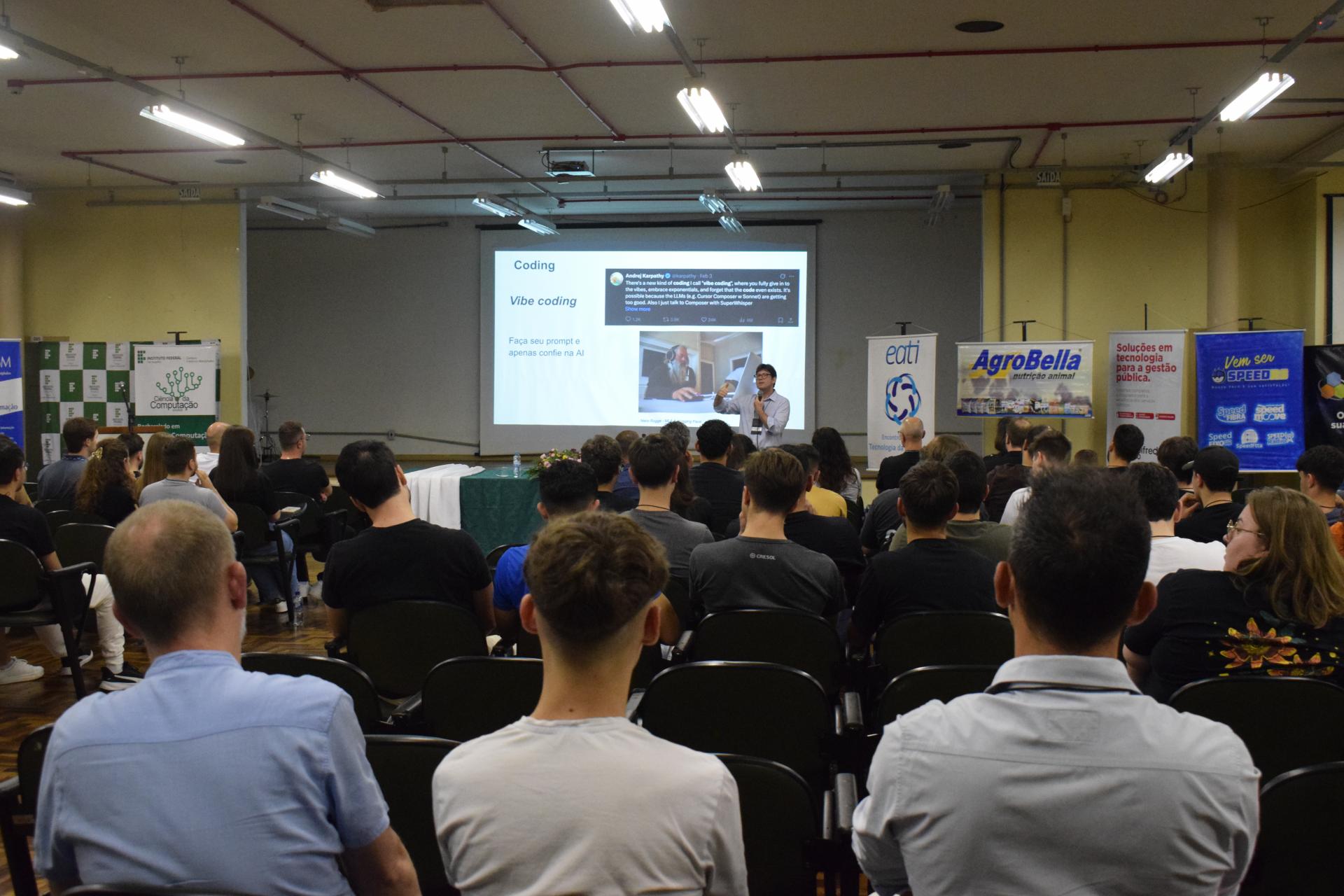 Foto colorida horizontal de palestra. Bem ao fundo, o palestrante. À frente dele um grupo expressivo de pesssoas sentadas. 