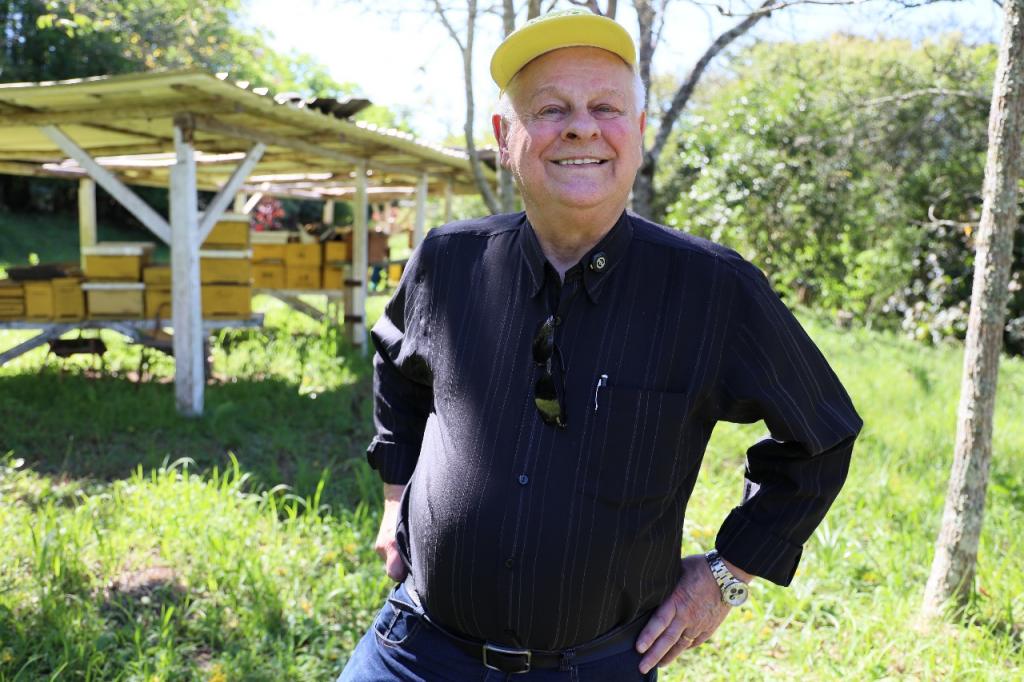 Foto colorida horizontal de homem idoso com camisa preta e boné amarelo. Ele está um local arborizado com um prédio de um andar ao fundo.