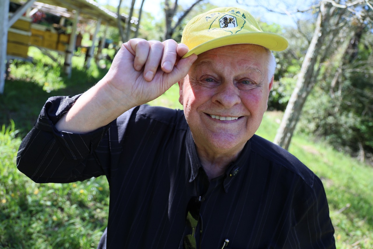 Foto colorida horizontal do professor Silvio com camisa preta segurando um boné amarelo com uma abelha desenhada. Ao fundo, área verde