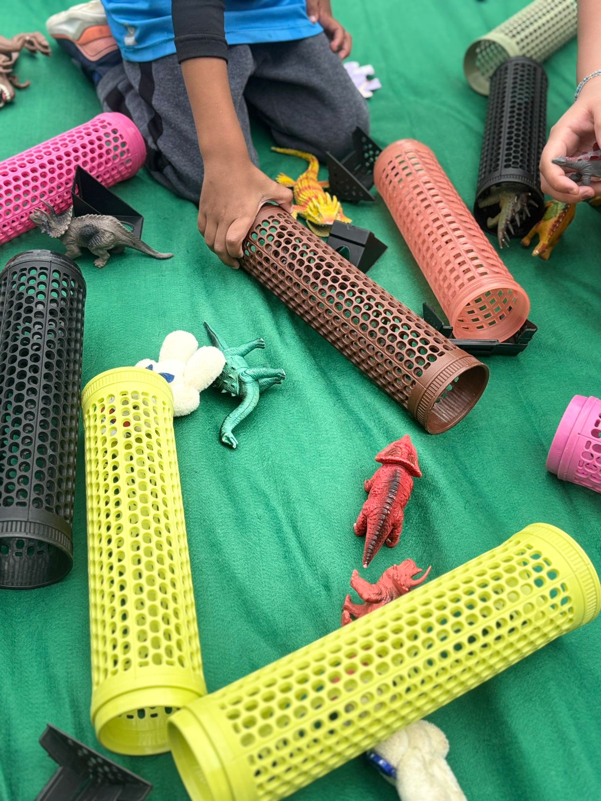 Foto colorida horizontal de canos coloridos de PVC e brinquedos em formato de dinossauros em cima de um pano verde. A imagem mostra a mão de uma criança manipulando um dos itens