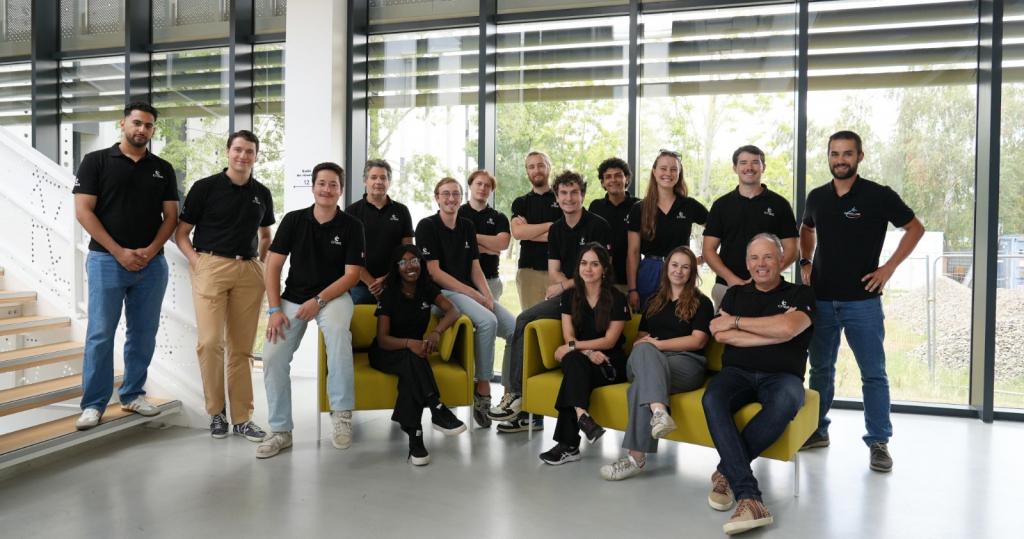 Duas fileiras de pessoas. As pessoas da fileira da frente estão sentadas. Todos utilizam camiseta preta com a logo da Agência Espacial Francesa.