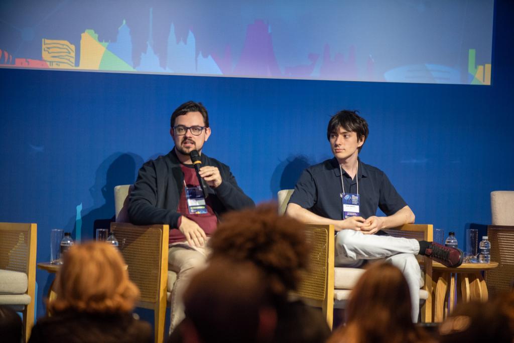 Foto colorida horizontal de dois homens jovens sentados em poltronas à frente de uma parede azul. O jovem da direita é o estudante Ricardo. À esquerda dele, outro homem segura o microfone. À frente deles, a plateia.