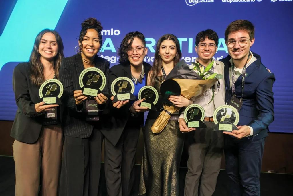 Foto colorida horizontal de seis jovens segurando troféus em formatos circulares. O grupo é composto por quatro mulheres e dois homens. Todos usam roupas sociais. Atrás deles um painel azul com a marca do evento