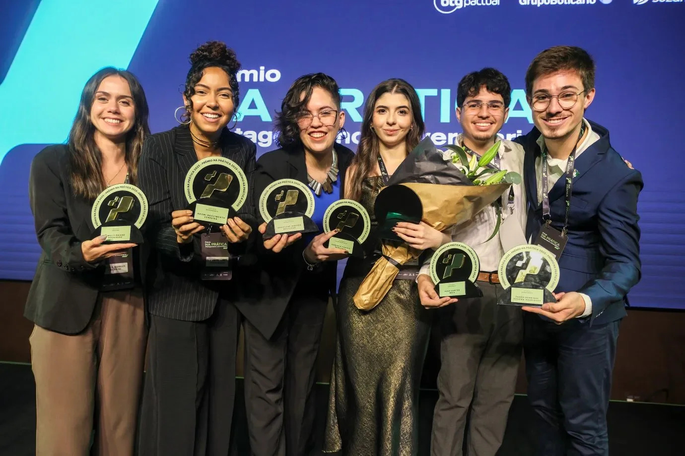 Foto colorida horizontal de seis jovens segurando troféus em formatos circulares. O grupo é composto por quatro mulheres e dois homens. Todos usam roupas sociais. Atrás deles um painel azul com a marca do evento