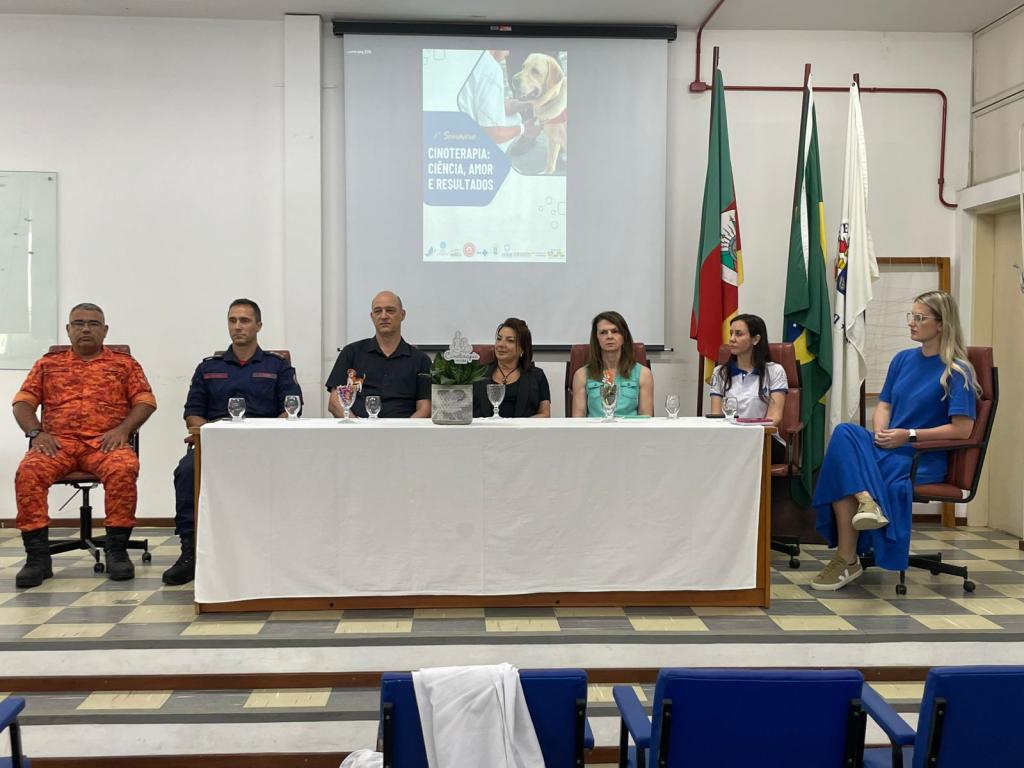 Foto horizontal e colorida de uma mesa de abertura com sete pessoas sentadas em um auditório, diante de um telão com a arte do evento. Bandeiras estão posicionadas ao fundo e o ambiente é organizado para uma apresentação formal.