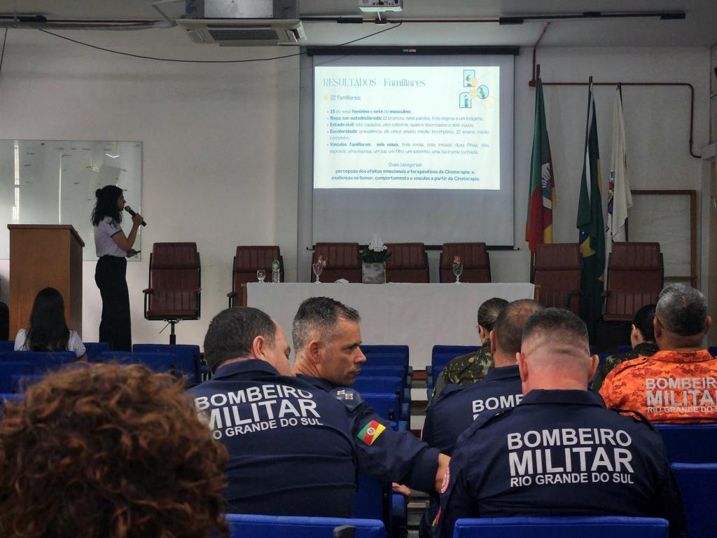 Foto horizontal e colorida de um auditório. À esquerda, uma mulher está em pé ao lado de um púlpito, segurando um microfone enquanto apresenta slides projetados na tela ao fundo
