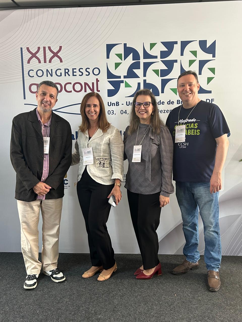 foto colorida vertical de quatro pessoas em pé, dos lados dois homens e no centro duas mulheres. Eles estão sorrindo e usam crachás brancos no peito. Ao fundo um banner branco do congresso