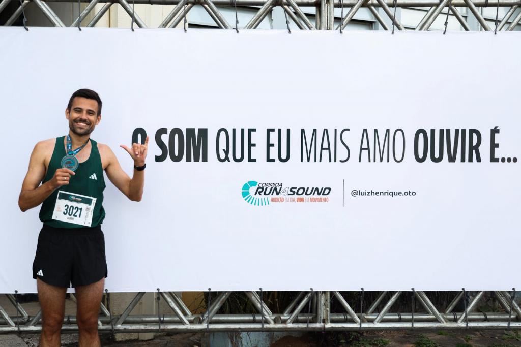 foto colorida horizontal de um homem segurando uma medalha pendurada no pescoço com uma mãe e fazendo um sinal com a outra, tendo ao fundo um painel branco da corrida