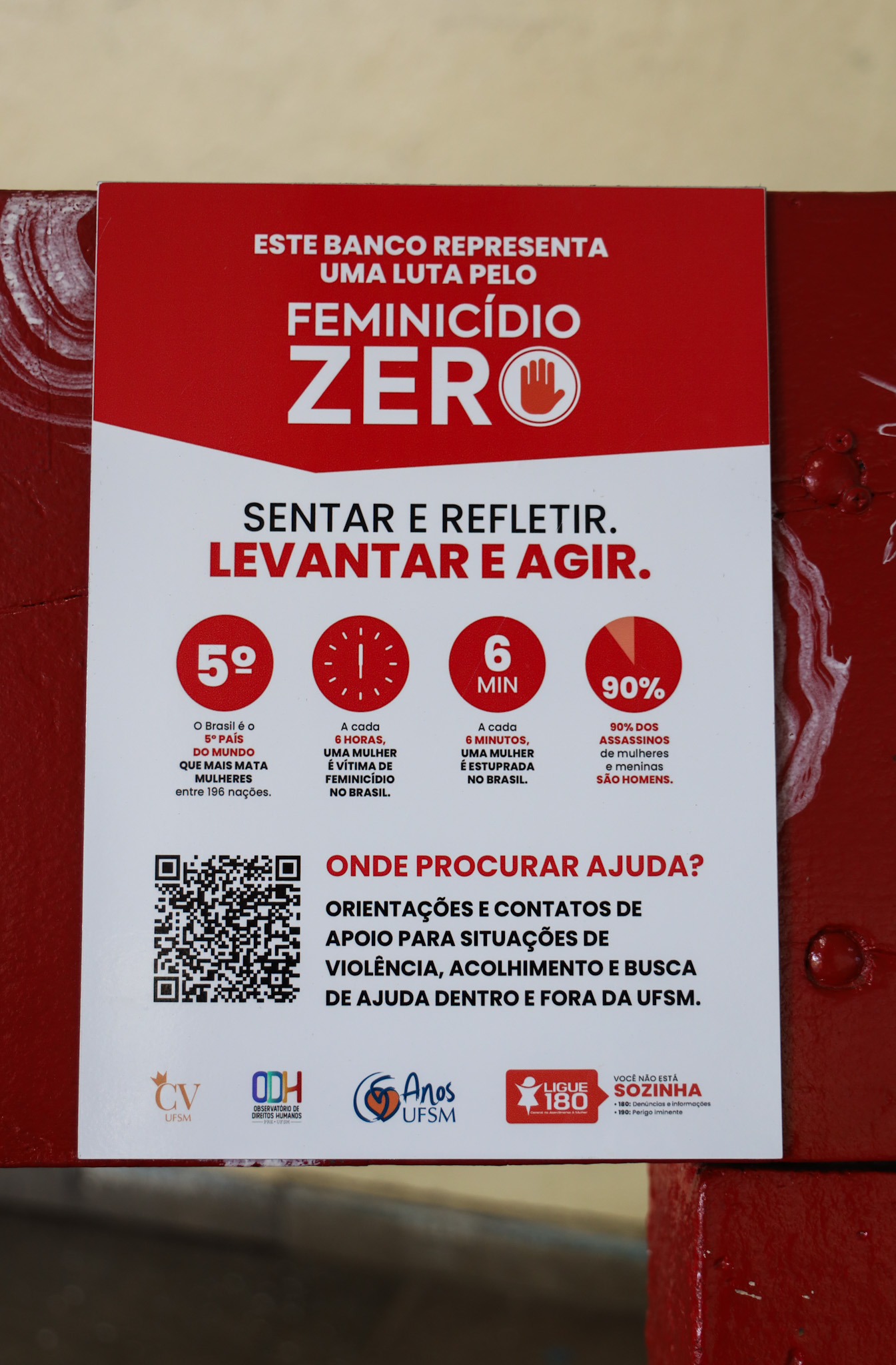 A fotografia é vertical e colorida. O foco da imagem é uma placa que está no baco vermelho. Na placa está escrito "este banco representa uma luta pelo feminicídio zero". Também mostra como procurar ajuda em casos de violência contra as mulheres.