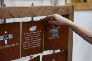 A foto mostra um jogo de madeira, que possui vários quadrados para serem girados. De um lado, há um termo da arqueologia e, do outro lado, a explicação do conceito. Na foto é possível ver a parte de cima do jogo, com três quadrados, e a mão de uma pessoa, branca, com unhas pintadas de preto, mexendo no segundo quadrado.