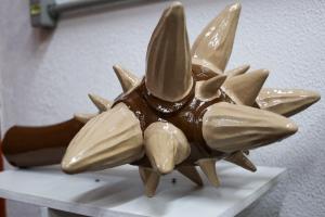 A imagem mostra uma réplica da cauda de um gliptodonte, uma espécie de tatu gigante. A cauda é comprida, marrom, e termina em uma forma arredondada, com protuberâncias no formato de espinhos, de cor bege, saindo dela.