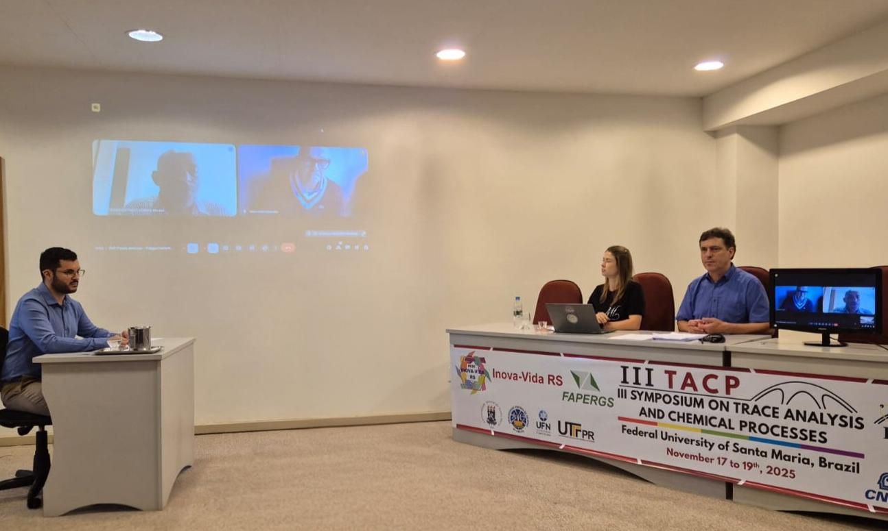 foto colorida horizontal de uma banca de defesa, com um homem sentado em uma mesa à esquerda, a banca, com duas pessoas, em outra mesa à direita, e na parede de fundo duas telas projetadas, cada uma com um homem