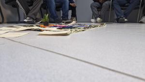 Foto horizontal. Mostra uma sala com chão e paredes na cor branca. Ao centro, há um espaço retangular sobre o qual estão depositados papéis e recortes de diferentes cores, com uma pequena planta dentro de um vaso. Seis homens estão sentados em cadeiras encostadas nas paredes. Na imagem, aparecem somente as suas pernas.