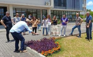 Foto horizontal. Tendo um prédio ao fundo, um homem agachado sobre um canteiro de flores fala para um público formado por cerca de 15 pessoas.