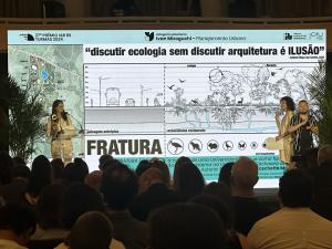 Foto horizontal. Uma plateia assiste à cerimônia de premiação, realizado sobre um palco, no qual há uma mulher no canto esquerdo (a qual segura um troféu e fala ao microfone) e, no canto direito, uma mulher que segura papéis e um rapaz que aparentemente faz tradução para Língua Brasileira de Sinais. Ao fundo, é projetado o trabalho premiado.