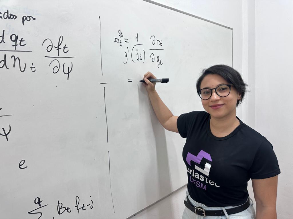 Fotografia colorida horizontal. A direita está Renata. Uma mulher de pele clara, cabelo escuro e curso. Ela veste uma camiseta preta. Ela escreve formulas científicas em um quadro branco