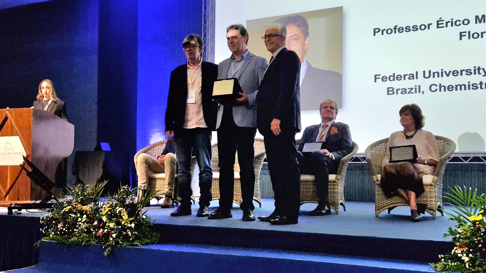 Foto colorida horizontal com três homens em pé. O que está ao centro é o professor Érico, que segura uma placa. Todos vestem ternos. 