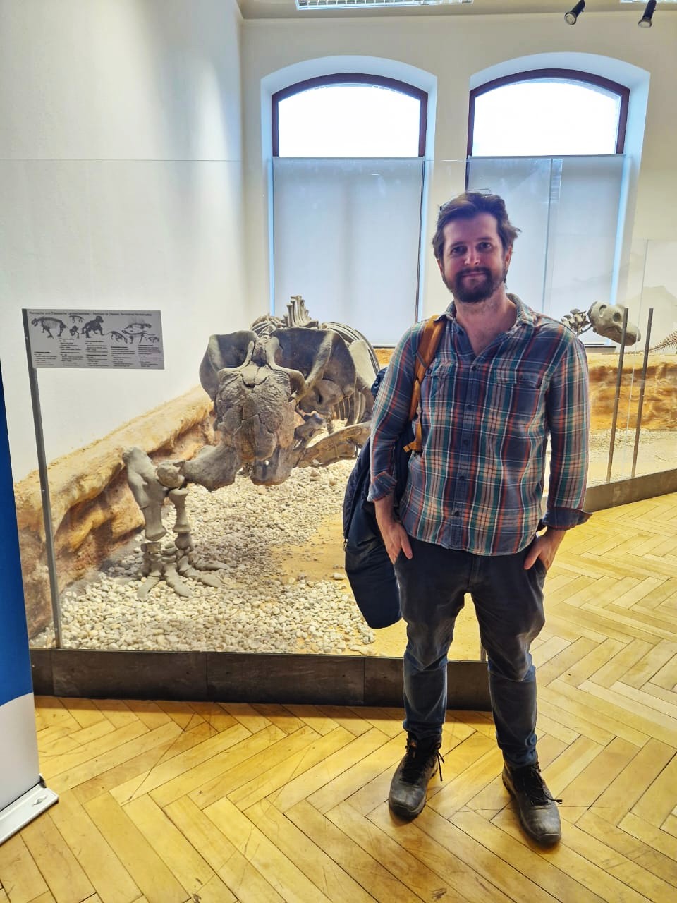 foto vertical colorida de um homem, de barba, camisa xadrez, mãos nos bolsos, à frente de uma estrutura de vidro com fósseis. O ambiente é uma sala iluminada, com duas portas de vidro ao fundo