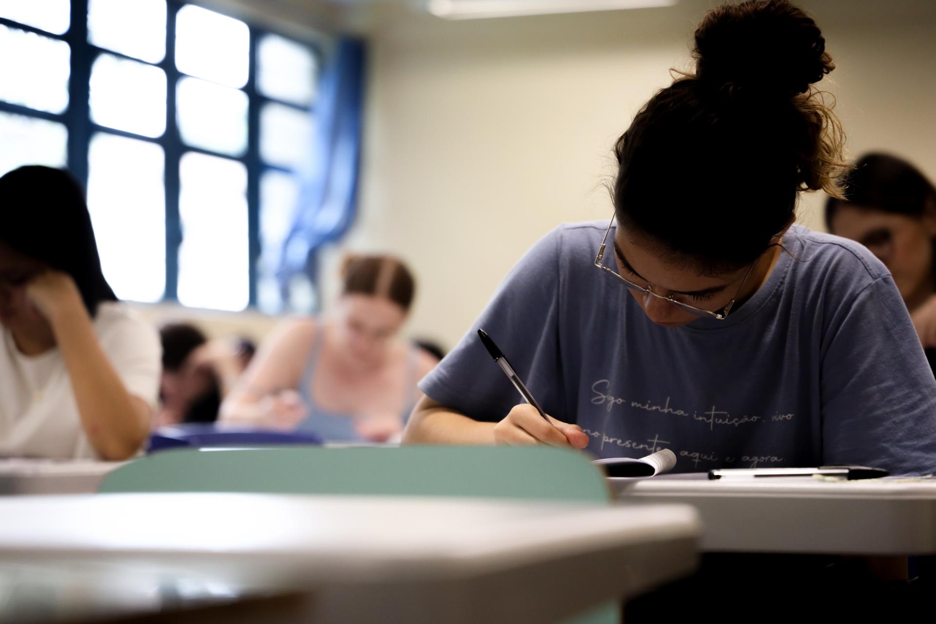 Foto horizontal, que mostra pessoas sentadas dentro de uma sala fazendo a prova
