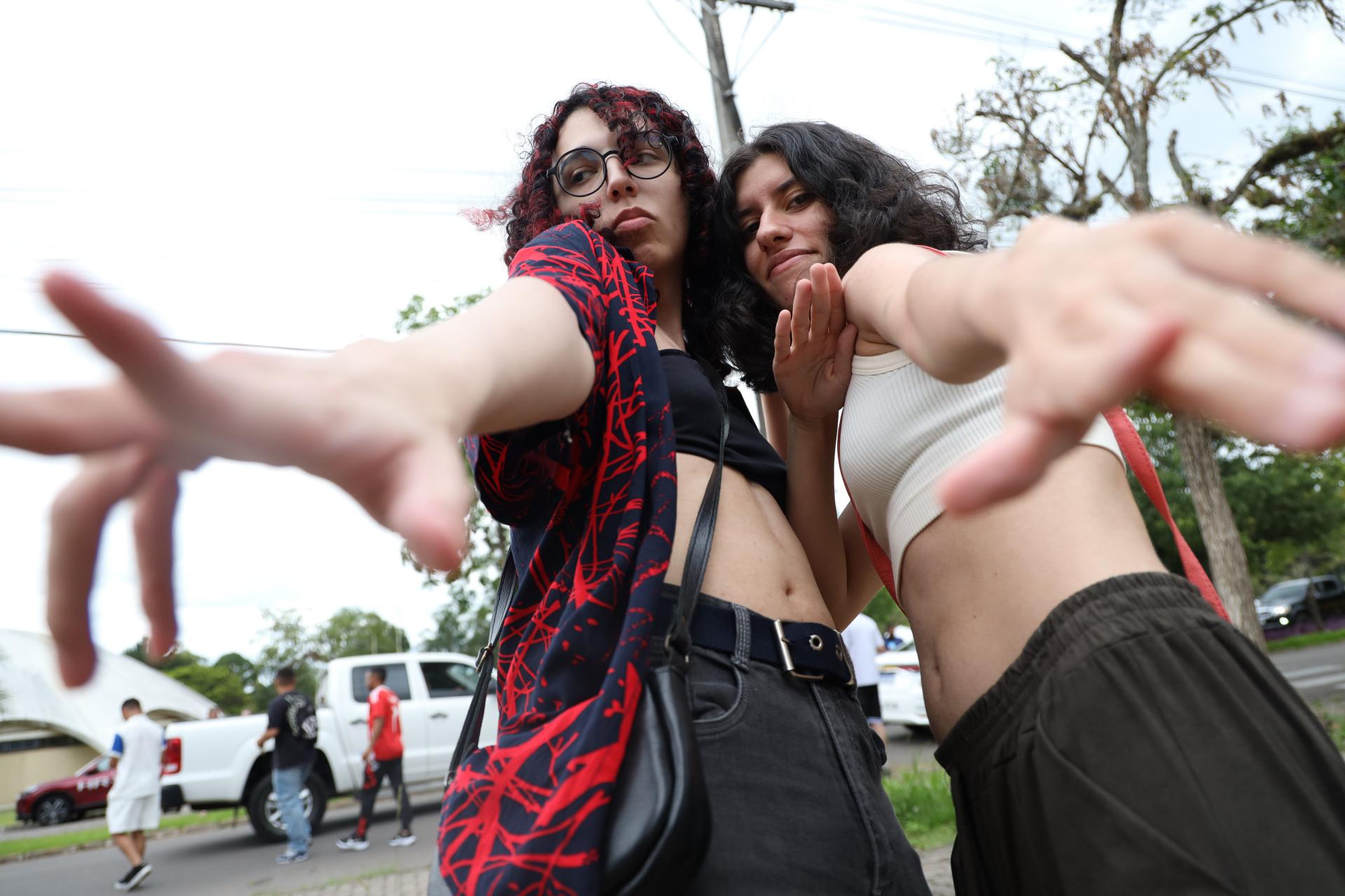 Foto horizontal. Duas meninas posam para foto depois de terminada a primeira prova do Vestibular. Ambas são brancas, têm cabelo escuro comprido e vestem calças pretas. Uma delas veste uma blusa preta e a outra, uma blusa branca.