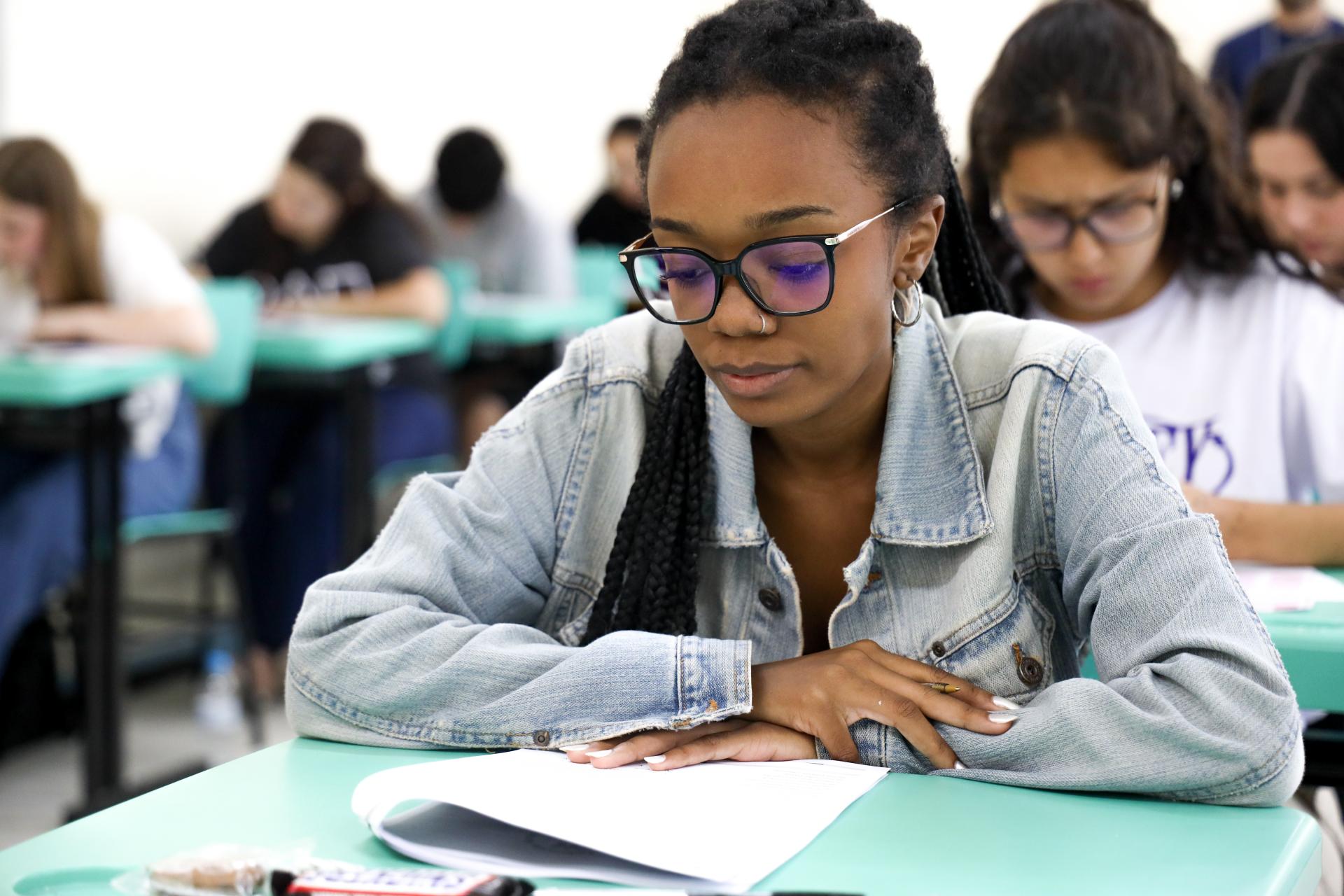 Foto horizontal e colorida de uma estudante negra realizando a prova do Vestibular em uma sala com outros alunos