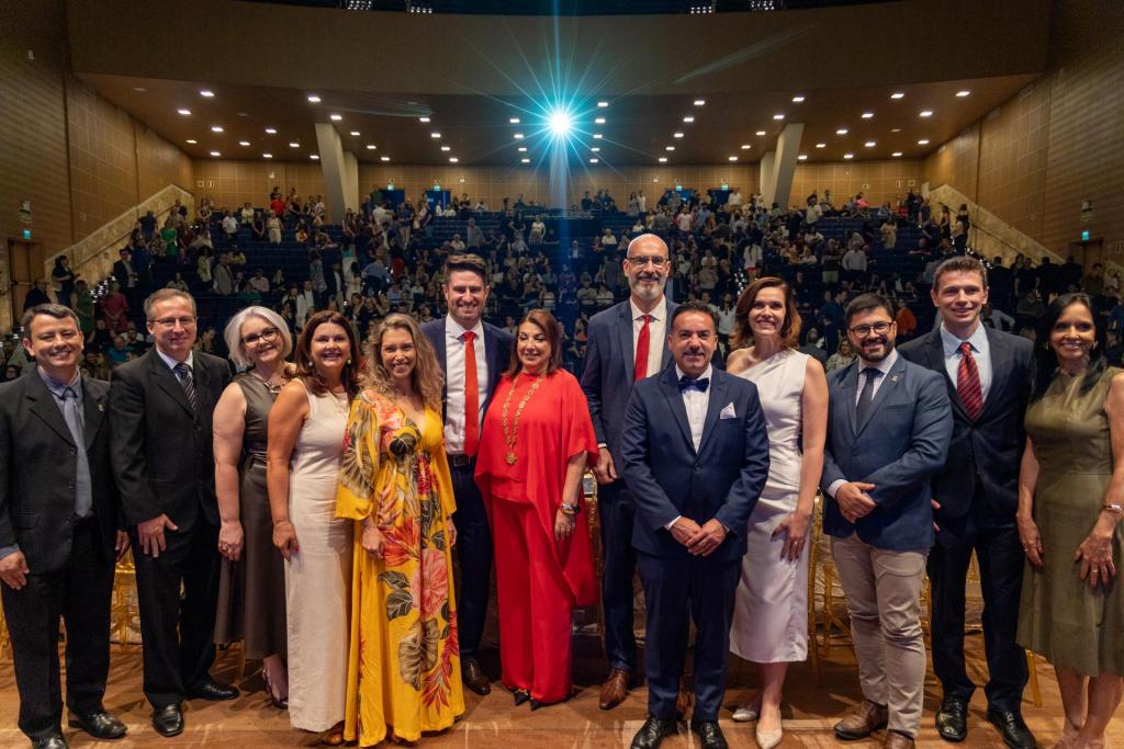 Foto horizontal, colorida e posada de Martha Adaime e Tiago Marchesan junto a equipe de pró-reitores e chefia de gabinete. Ao fundo, está a plateia do Centro de Convenções da UFSM