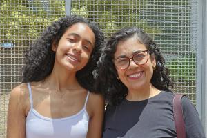 Foto horizontal, que mostra mãe (à dir.) e filha (à esq.). Ambas têm pele morena e cabelo comprido e crespo. A mãe veste uma blusa preta e a filha, uma blusa branca.