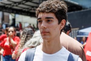 Foto horizontal: Mostra um rapaz branco com cabelo curto e castanho, que veste uma camiseta branca e tem dois brincos na orelha esquerda, além de estar com uma mochila nas costas.
