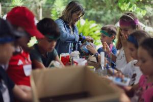 Várias crianças confeccionam brinquedos com materiais recicláveis, uma mulher de cabelo loiro e jaqueta jeans as ajuda.