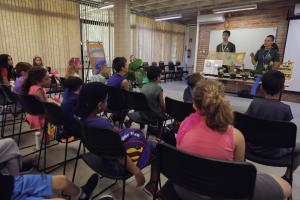 Em um auditório, várias crianças estão sentadas de costas em cadeiras pretas, observando dois adultos de frente para elas, usando camisetas verdes e mostrando plantas em uma mesa.