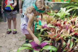 Uma menina loira, usando roupas roxas claras e com uma faixa verde na cabeça, está olhando para baixo e tocando uma planta.