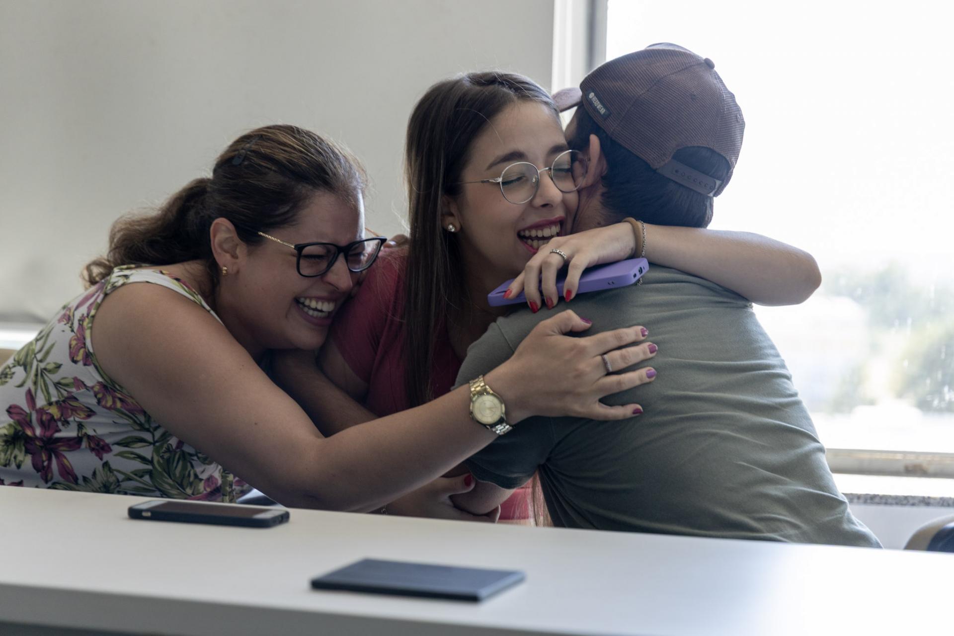 Foto horizontal e colorida de mãe, filha e pai se abraçando e comemorando sentados.