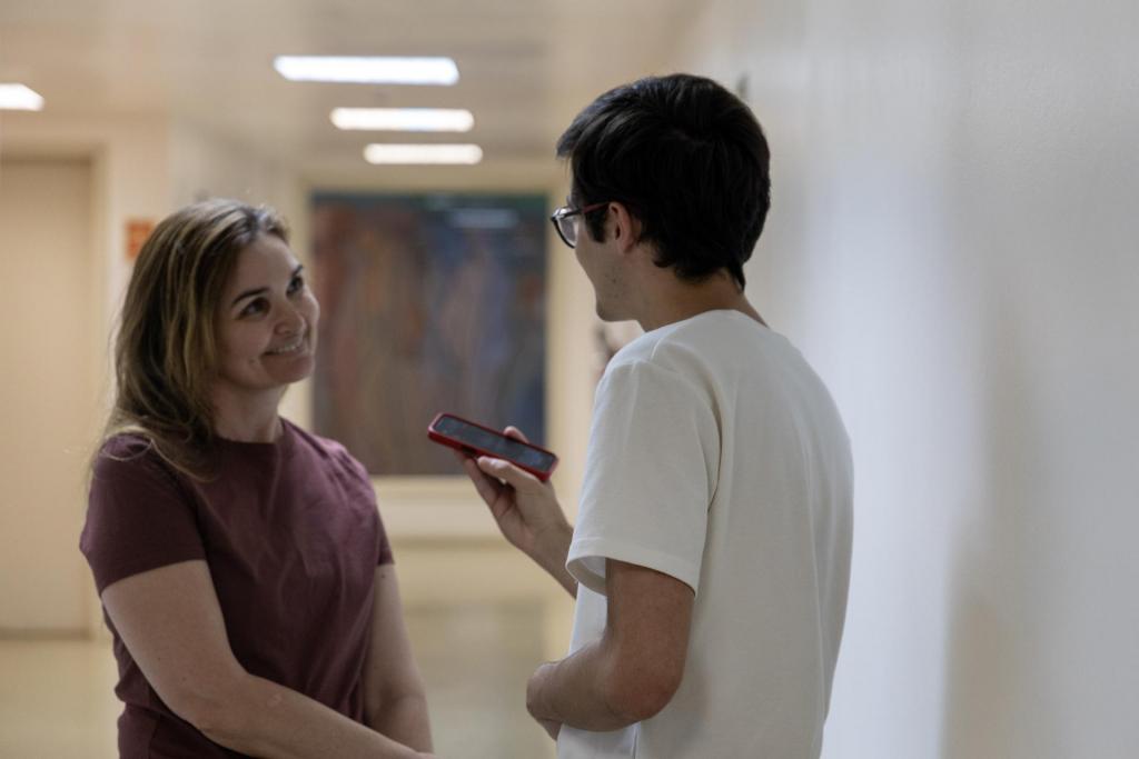 oto horizontal e colorida de uma mulher sendo entrevistada por um homem. Na imagem, a mulher está à esquerda e de olhando para o repórter. Já o repórter está de costas para a câmera, no lado direito, e segura um celular como microfone