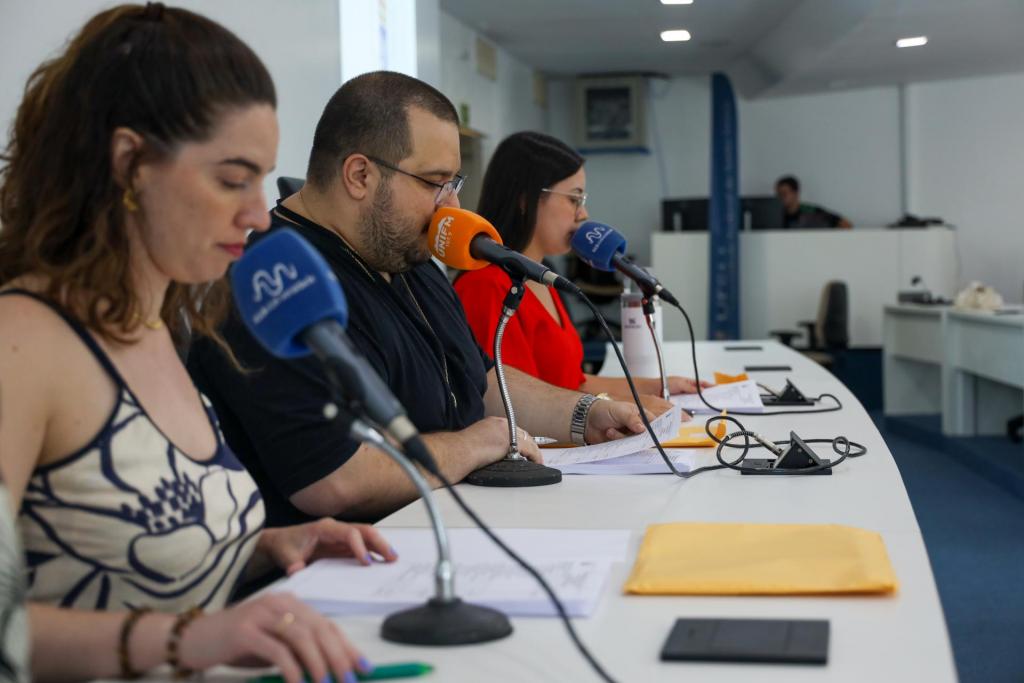 Foto horizontal e colorida de duas mulheres e um homem sentados lendo os nome dos aprovados em microfones.