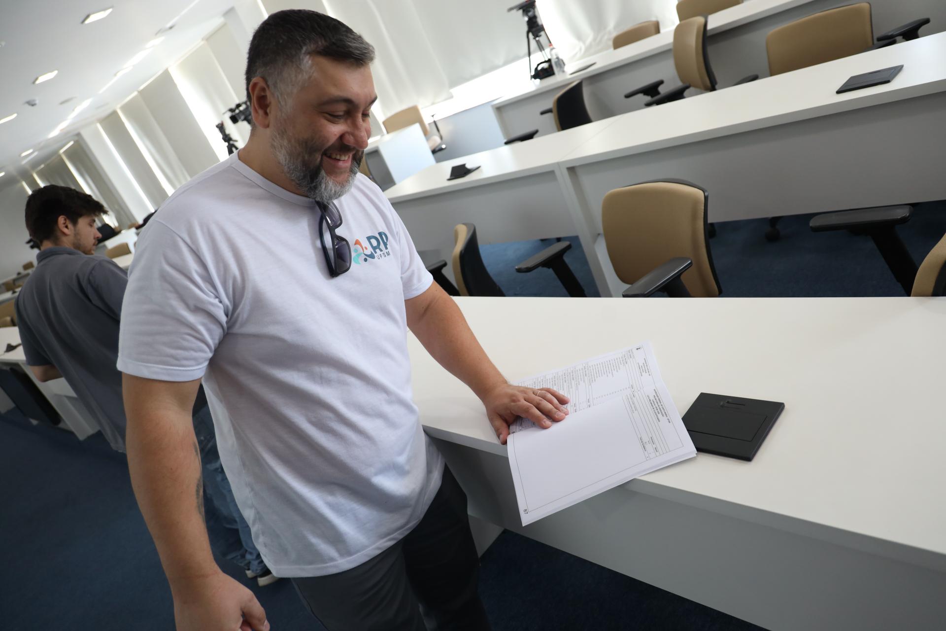 Foto horizontal e colorida de um homem em pé sorrindo enquanto lê a lista de aprovados.