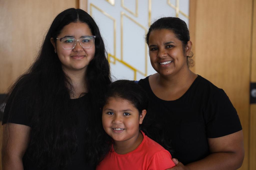 Foto colorida horizontal de família composta por filha adolescente, filha criança e mãe. A filha mais velha usa óculos, tem o cabelo solto e longo, e está à esquerda da mãe. A menor tem cabelo preso e está à frente da irmã e da mãe, que também está de cabelo preso. A imagem mostra as três da cintura para cima