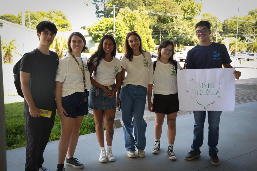 Foto colorida horizontal de seis pessoas em pé, sendo dois homens e quatros mulheres jovens. Eles seguram um cartaz Bixos Biologia