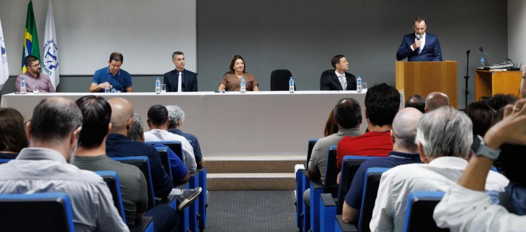 foto colorida horizontal de pessoas sentadas em uma mesa oficial, de frente para o público, que aparece de costas