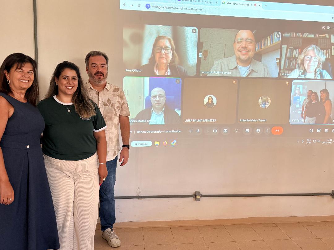 Foto colorida horizontal de três pessoas em pé posando ao lado de uma chamada em vídeo com pelo menos cinco pessoas. As pessoas em pé são duas mulheres, uma com vestido preto, outra com blusa preta e calça branca, e um homem com calça jean e uma camisa estampada