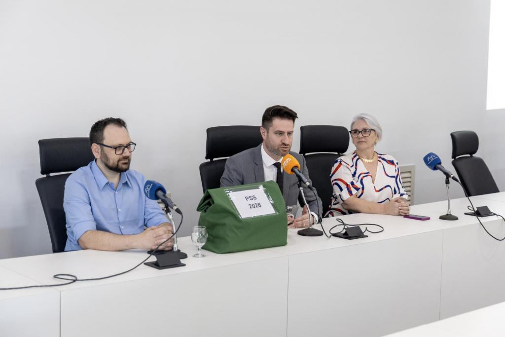 Foto horizontal e colorida de dois homens e uma mulher em uma mesa branca. Eles vestem roupas formais. Microfones e o malote com a lista de aprovados estão na mesa.