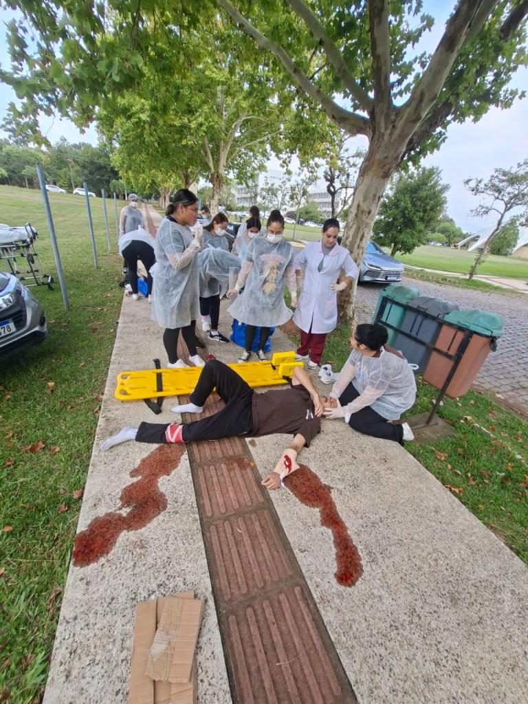 Um homem branco descalço, vestindo meias brancas, calças pretas e camiseta marrom está deitado sobre uma calçada, entre um gramado e uma rua de paralelepípedos. Ele está enfaixado na perna esquerda e no braço direito, membros dos quais simula-se um sangramento, com líquido vermelho jogado sobre a calçada. Ao lado dele, há uma maca amarela. E seis mulheres vestidas de branco,  estão em volta dele, simulando o atendimento a uma vítima.