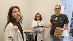 Fotografia horizontal colorida com três pessoas enquadradas, uma ao lado da outra, sendo duas mulheres e e um homem. Na imagem, a mulher ao centro segura o braço da outra e, ao mesmo tempo, entrega um tubo com sangue coletado