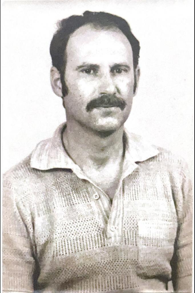 Foto em tons de cinza vertical de um homem adulto com cabelo escuro e curto, com bigode. Ele usa uma camisa social de manga longa com bolso de cor clara.